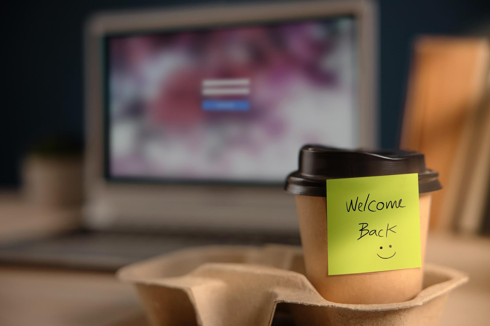coffee cup on a worker's desk with a sticky note that reads welcome back