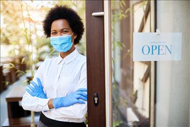 Business owner wearing mask proudly standing in front of her open restaurant