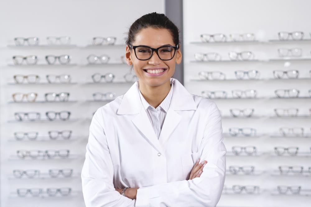 Happy optometrist posing in front of glasses on shelves