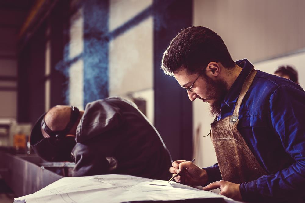 welding team member checking blueprints while another welder works in the background