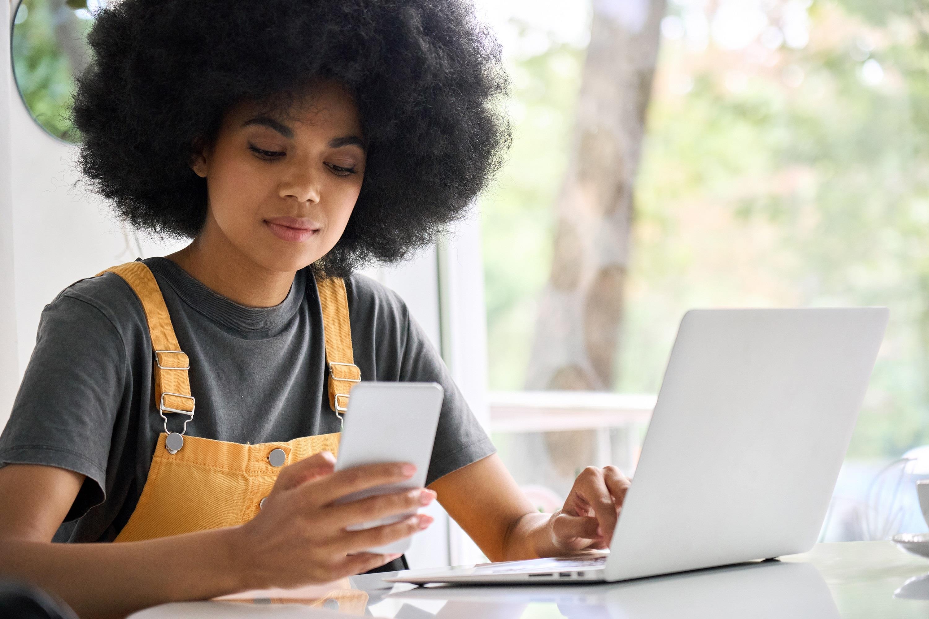 gen z job seeker looking at her phone while researching jobs on her laptop