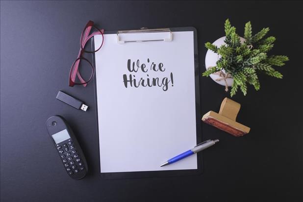 "we're hiring" written on a clipboard with other items on a desk