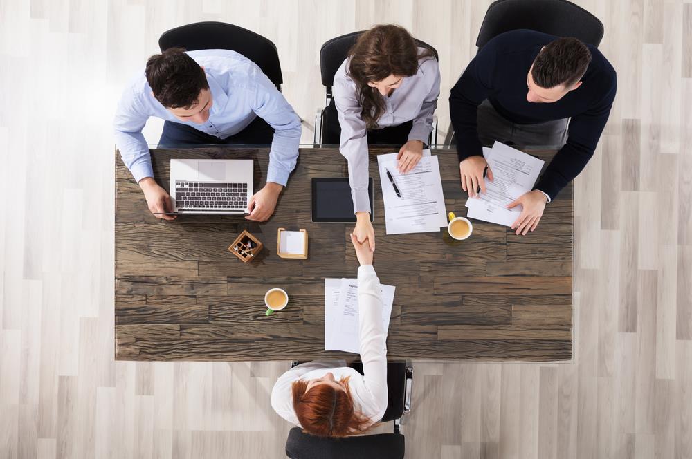 One interviewer from a panel of three shakes the hand of the interviewee across the table